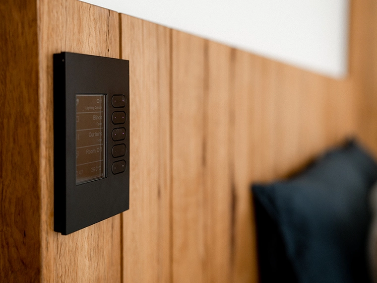 Wall‑mounted black smart control panel with a small display and buttons on a wooden interior wall.