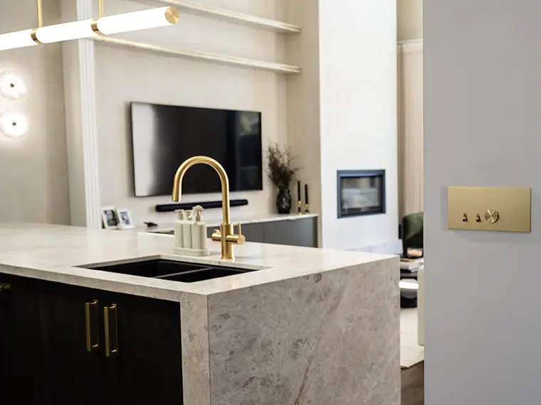 Brushed brass Solis switch on wall in kitchen with marble counter and gold faucet.