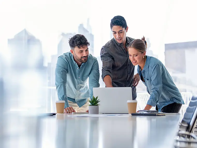 Three colleagues collaborating around a laptop in a bright office with coffee cups.