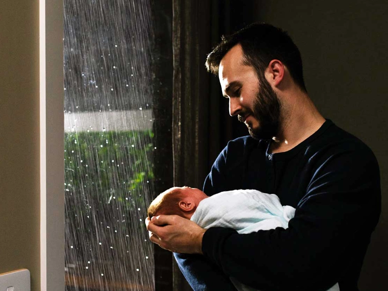 A man holding a sleeping baby by a window overlooking rainy weather outside.