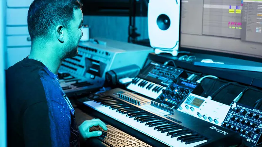 Guy Sebastian at his in-home studio, working on music with a large monitor, keyboards, synthesizers, and audio gear.
