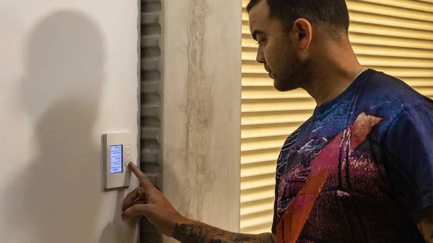 Guy Sebastian interacting with a wall-mounted C-Bus control panel, next to a window with closed blinds
