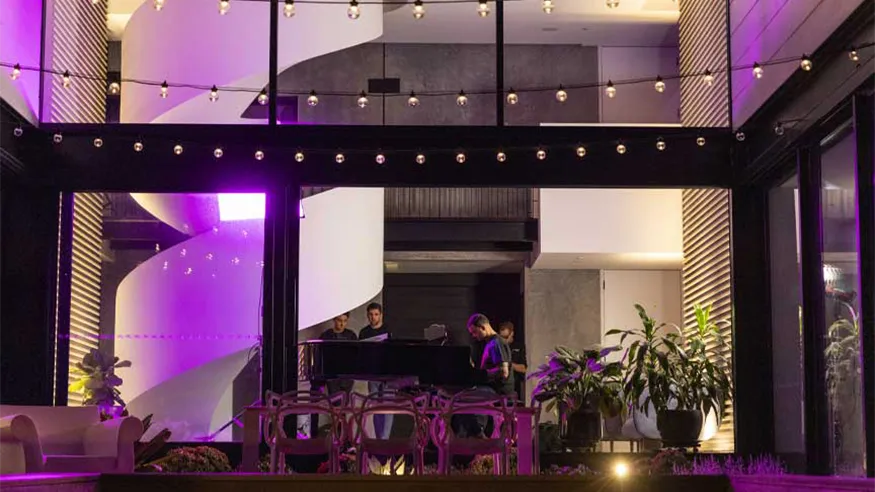 Modern backyard with string lights, plants, and seating; indoors, a spiral staircase and people around a grand piano.