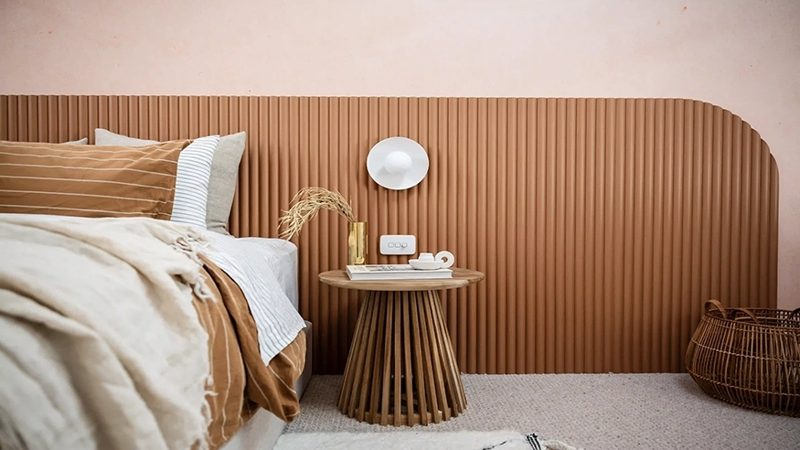 Modern bedroom featuring a stylish home renovation with a wooden slatted headboard, neutral-toned bedding, and decorative accents on a round side table with an Iconic Essence cover plate above the side table.