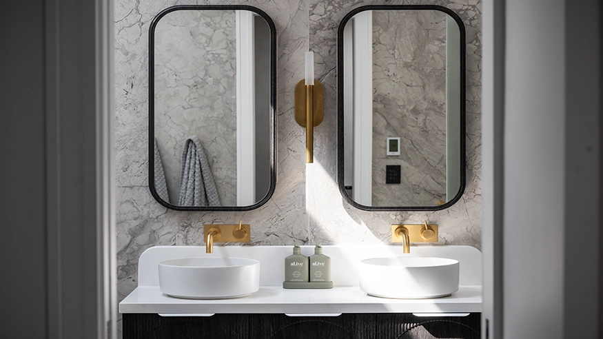 Modern bathroom with marble walls, black-framed mirrors, gold faucets, and a central light fixture, highlighting a stylish home renovation. A black Iconic Styl can be seen through one of the mirrors.