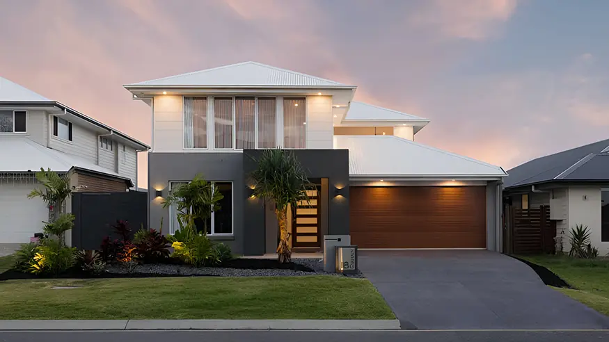 Modern two-story house with white roof, large windows, wooden garage door, and landscaped front yard at sunset.