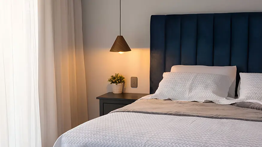Modern bedroom featuring Iconic Essence switch in Ash Grey, navy headboard, white bedding, and bedside table with pendant light.