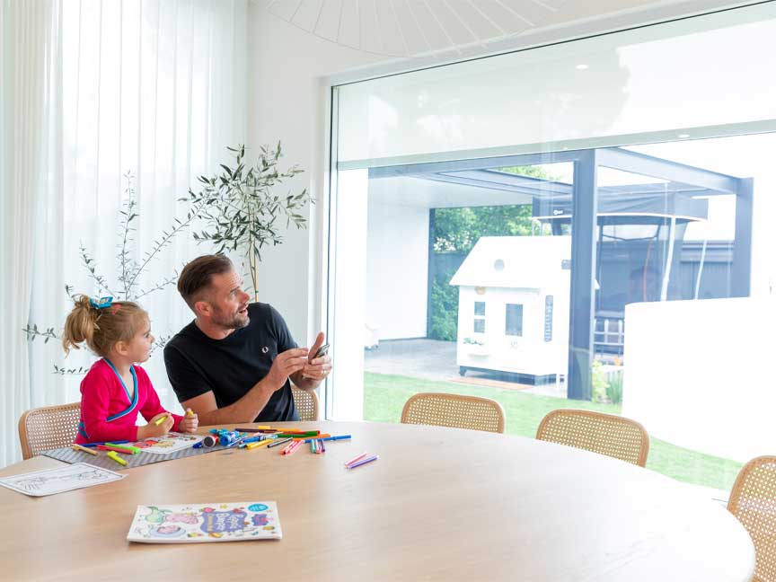 Troy Klemm and his child sitting at a round wooden table in a sunlit room with a large window. Klemm is on his smart phone controlling his smart blinds.