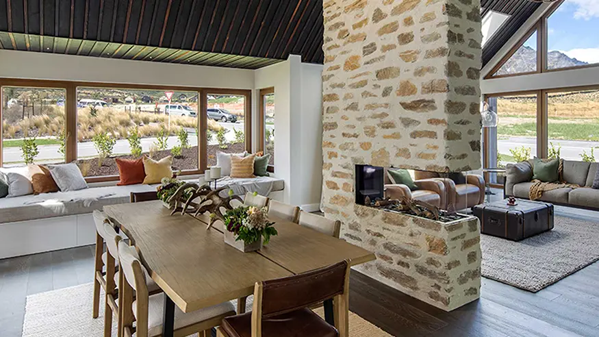 Open-plan dining and living area with stone fireplace, wood table, and large windows overlooking scenic views.