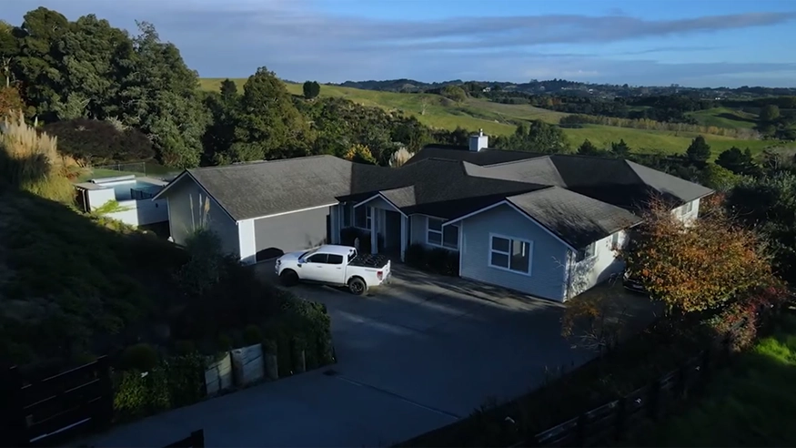Exterior view of a large rural Auckland home with gray roof, white walls, driveway, and surrounding greenery.