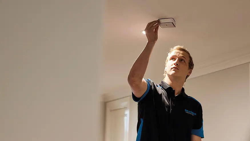 Electrician in dark shirt adjusts a PDL Fire Tek smoke alarm on the ceiling.