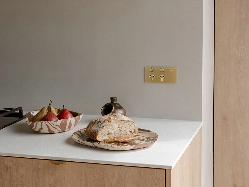 A white countertop displays a plate of bread and a bowl of fruit, while a Clipsal Solis wall-mounted switch and power points enhance the aesthetic design.