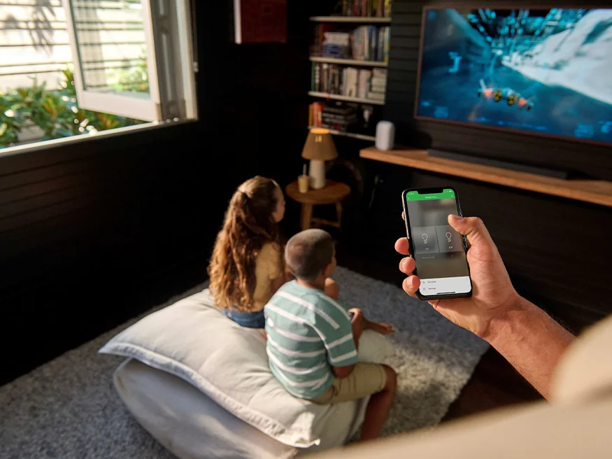 Person holding a smartphone controlling smart lighting while two children sit watching TV in a living room.