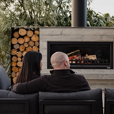 Two people sitting on an outdoor sofa facing a modern fireplace, with stacked firewood and greenery in the background.