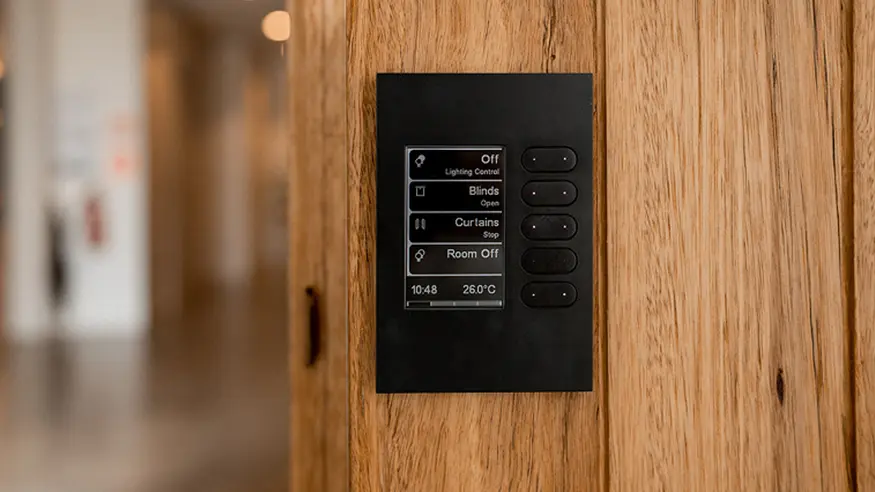 A C-Bus smart home control panel installed on a wooden wall, fetauring touchscreen options for lighting, blinds, curtains, and room shutdown.