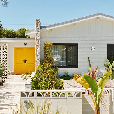White brick house with yellow double front door, black-framed windows, and tropical plants in the front yard.