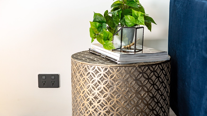 Stylish living room with a dark blue sofa, geometric side table, and a wall-mounted PDL Iconic Power point beside a stack of magazines and a potted plant.