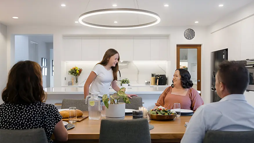 Modern kitchen and dining area with wooden table set for a meal, featuring salads, bread, and drinks.