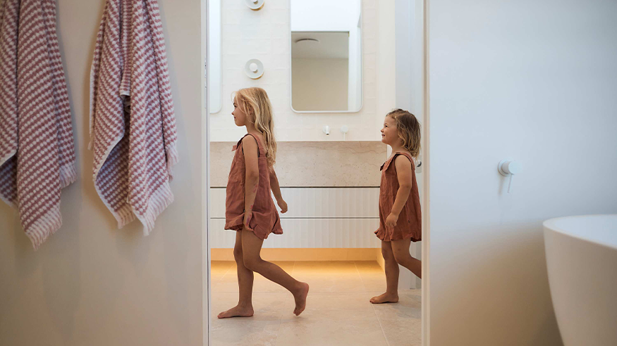 Two young girls walking together in a brightly lit bathroom, surrounded by colourful towels and bathroom fixtures.