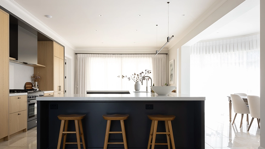 Modern kitchen with light wood cabinetry, large island and wooden stools, with natural lighting from sheet-curtained windows.
