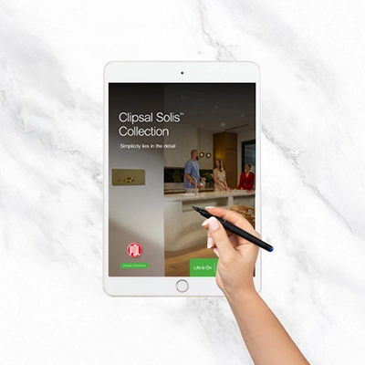 Hand holding a stylus over a tablet showing Clipsal Solis Collection brochure on a marble surface.
