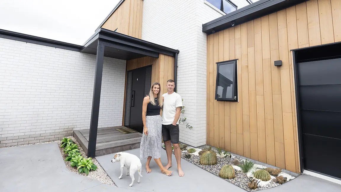A couple stand barefoot outside a modern house with white brick and wood paneling, next to a small white dog.