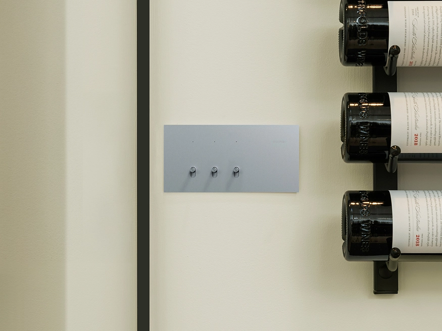 Clipsal Solis T Series 3-gang Flush Switch in Platinum Aluminium finish installed on a wall beside wine racks.