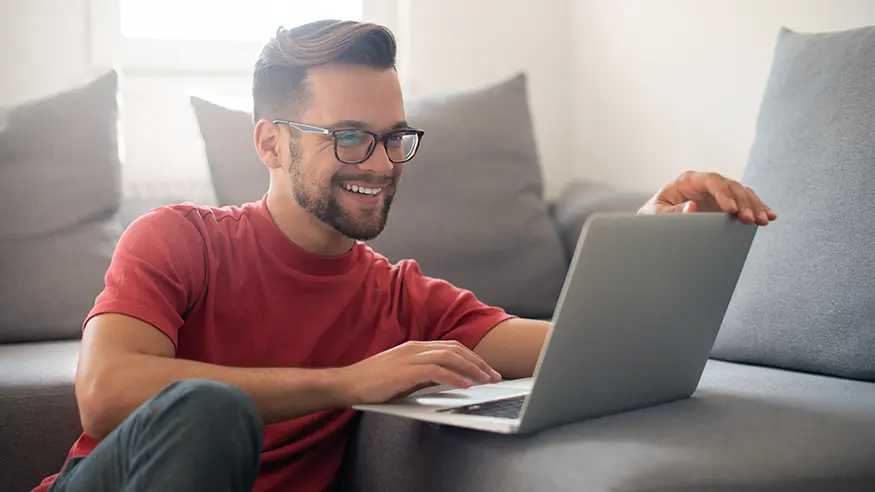 Person in red shirt using a laptop while seated on the floor beside a couch in a casual setting.