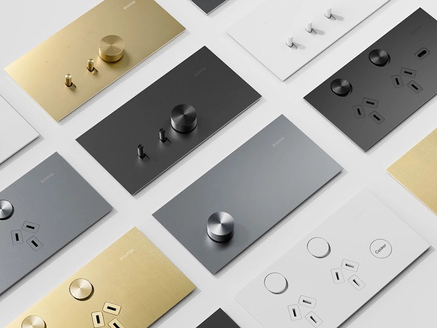 Clipsal Solis switch and power outlet collection in brushed brass, platinum aluminium, ebony aluminium, and white ceramic finishes arranged on a flat surface.