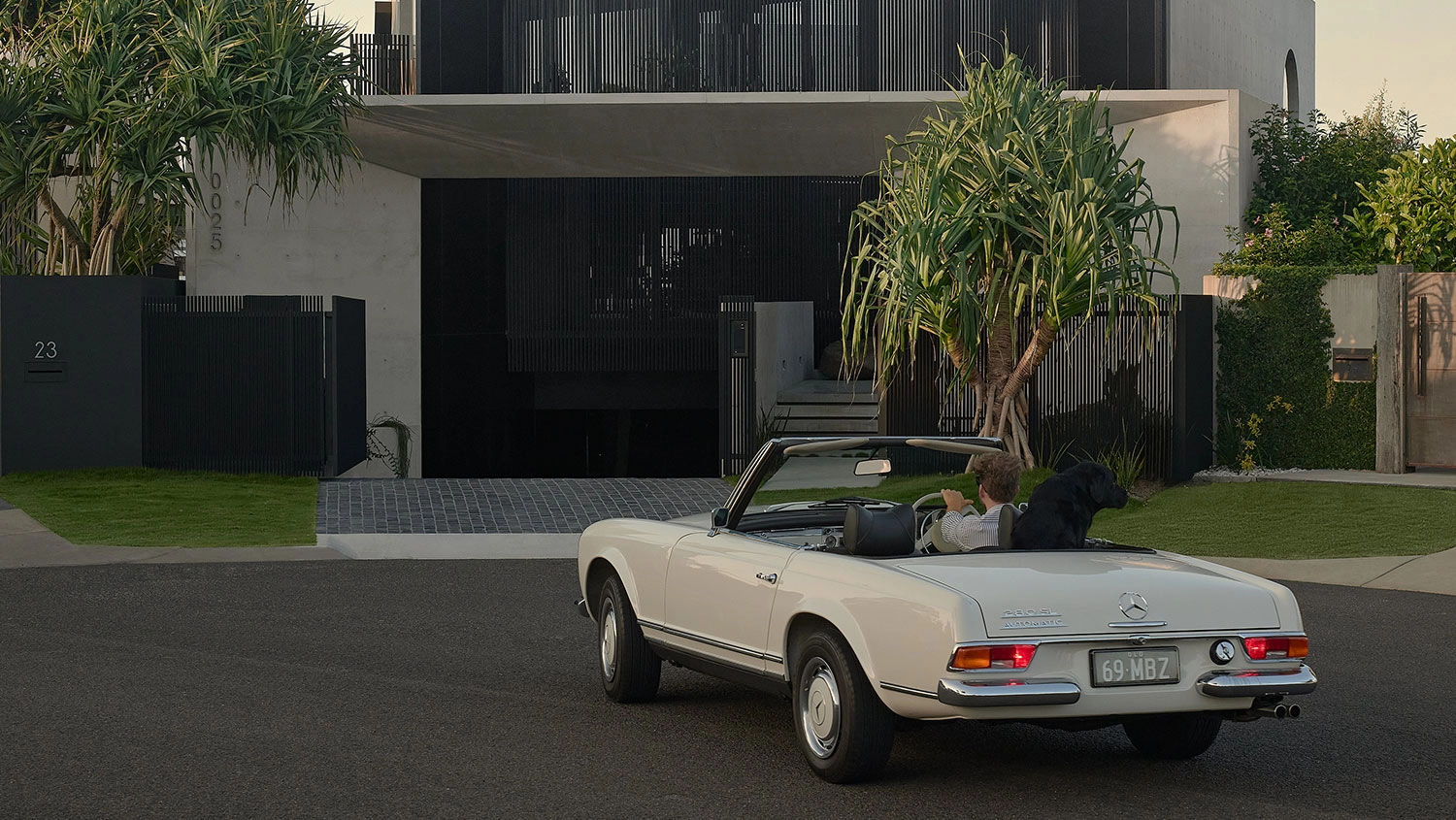 A young man is sat in the driver’s seat of a white convertible parked in front of a modern house with black gates. A large black dog standing with its front paws on the car door.