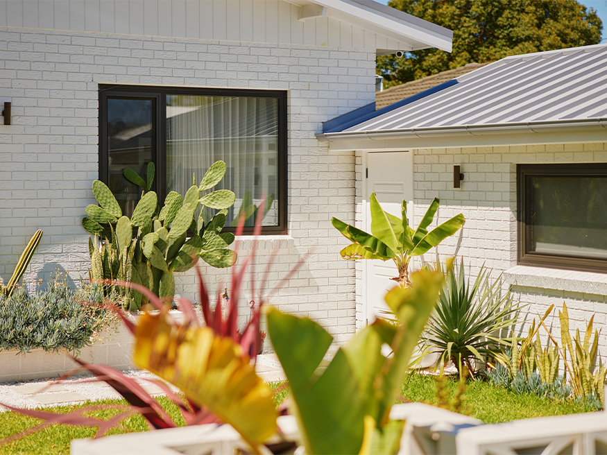 A white house with a roof and white fence, illuminated by daylight and bordered by plants.