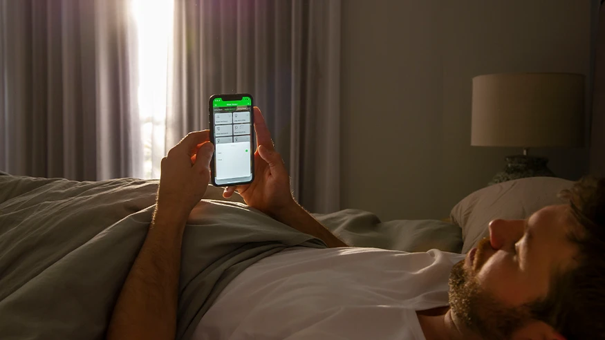 A man relaxing in bed, using his mobile phone to control smart home features via the PDL Wiser app.