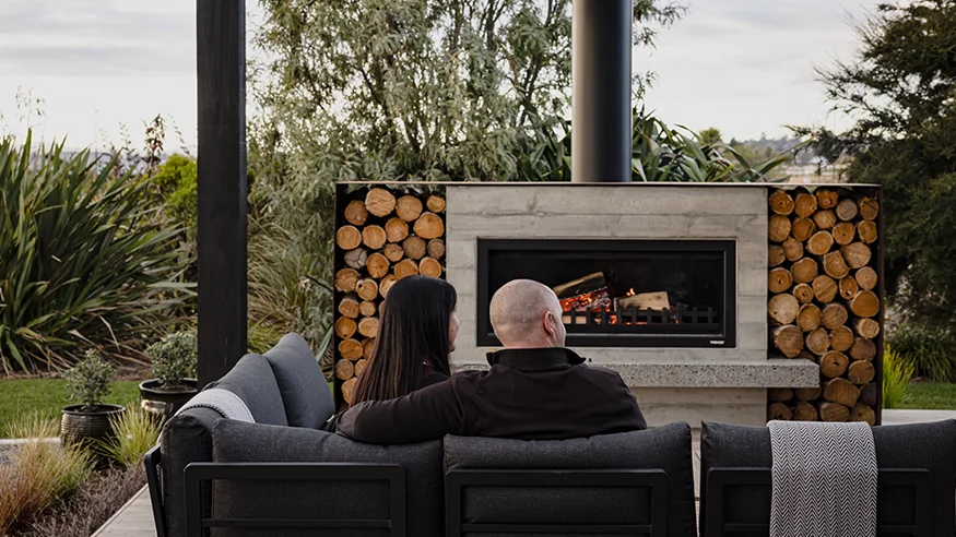 Two people sitting on an outdoor sofa facing a modern fireplace, with stacked firewood and greenery in the background.