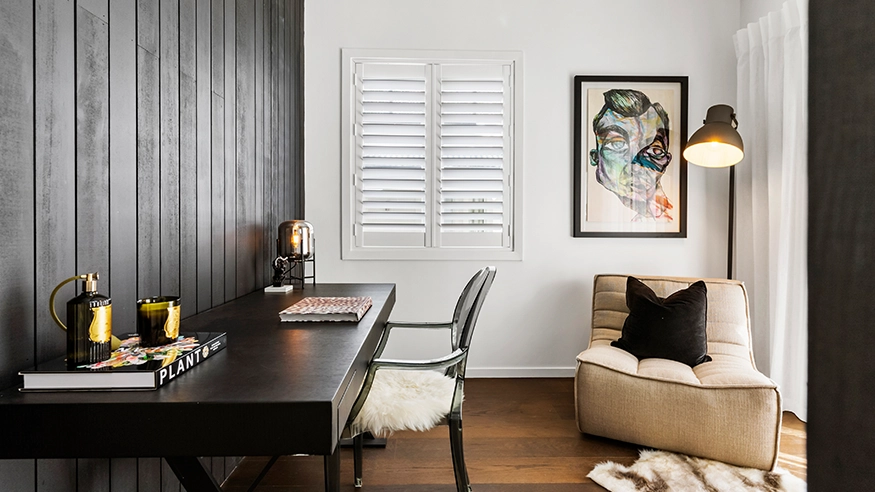 Modern home office with black desk, artwork, cosy lounge chair, and window shutters.